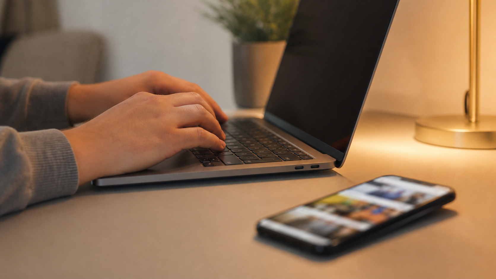 hands typing on laptop keyboard with a smartphone showing Instagram open on a desk